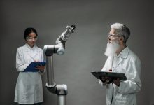Dois pesquisadores de jaleco trabalham com um braço robótico em um laboratório.