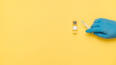 Mão enluvada segura uma seringa próxima a um frasco de vacina sobre fundo amarelo.