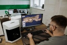 Profissional trabalha em um laboratório odontológico diante de um computador, criando um modelo digital de dentes.